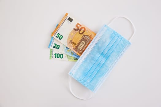 Surgical face mask covering cash on white background. Flat lay composition of euro bank notes hidden by medical equipment from directly above. Concept of budget cuts during coronavirus pandemic.