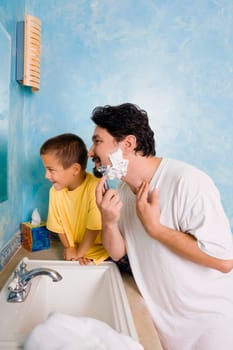 Side profile of a mid adult man shaving with his son sitting beside him in the bathroom