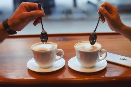 Two cups of coffee on the table, a man's hand and a woman's hand with spoons.