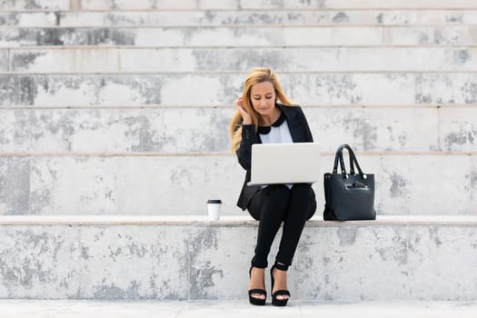 Young attractive businesswoman sitting on the stairs and using modern laptop