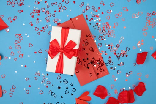 Presnet with envelope lying on blue background with glitters from directly above. Box with ribbon and red postcard on colorful desk in flat lay. Gift and card with heart sequins.