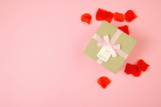 Pink background with present and rose leaves around. Golden box with ribbon from directly above. Gift lying on colorful table with copy space in flat lay composition.