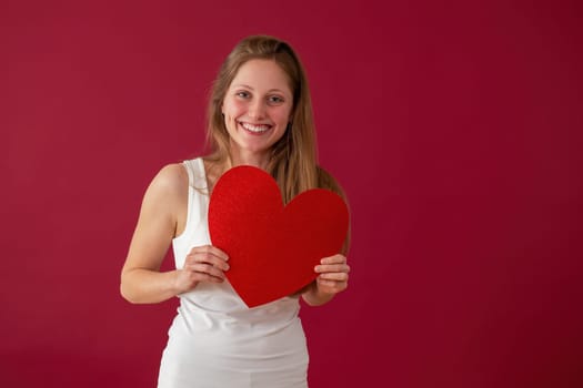 Smiling woman holding papper heart in hands on red background. Happy female with symbol of love on valentine's day. Joyful blonde girl with sparkling sign.