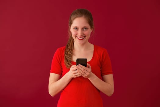 Blonde girl smiling with phone in hand on red background. Happy woman with lipstick texting on mobile on valentine. Positive female sending love message.