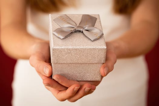Blurred female holding silver gift in hands on red background. Woman with little present with ribbon showing to the camera from close up.
