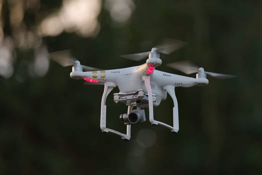 12 - White quad rotor drone with orientation lights hovering isolated from dark defocused background. Professional camera on gimbal beneath gives FPV
