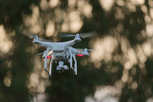 21 - Sunset bokeh background behind white quadcopter camera drone. Golden hour sun light creates dreamy smooth bokeh background shining through trees