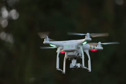23 - Green and red orientation lights on white quadcopter camera drone as it flies towards dark out of focus trees