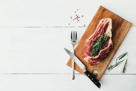 Raw ribeye steak on board, knife, fork, pepper, rosemary and pink Himalayan salt. High quality photo