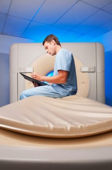 Healthcare worker sitting on a CAT scan machine and writing on a clipboard