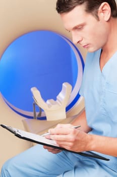 Healthcare worker sitting on a CAT scan machine and writing on a clipboard