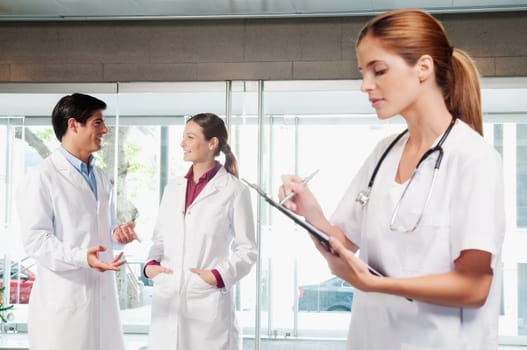Female nurse writing on a clipboard with her colleagues discussing in the background