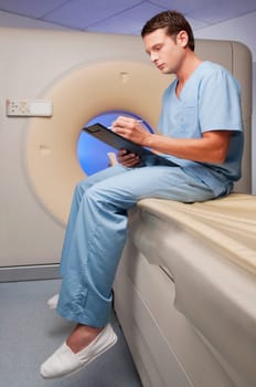 Healthcare worker sitting on a CAT scan machine and writing on a clipboard