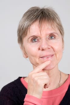 Head portrait of an elderly woman in front of a light gray background. Looking happily at the camera with her left index finger on her chin.