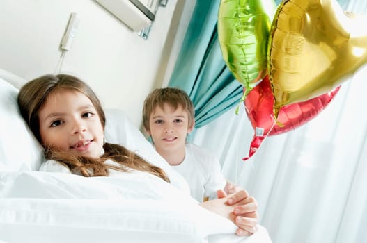 Girl holding balloons on a hospital bed with her brother beside her