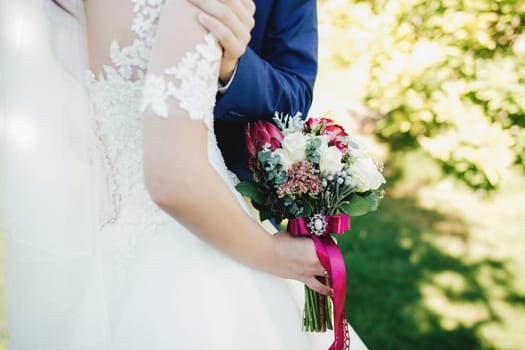 Bride in white dress is holding wedding bouquet. Groom hugs bride. Blurred background. High quality photo