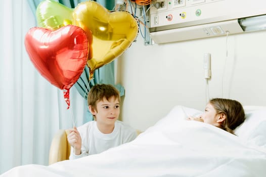 Girl on a hospital bed with her brother holding balloons