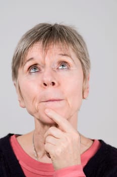 Head portrait of an older woman in front of a white background pensive and pressing lips, looking up with the left index finger on her chin.