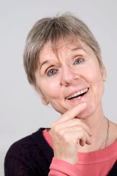 Head portrait of an older woman in front of a light gray background looking happily at the camera with her left index finger on her chin and open mouth.