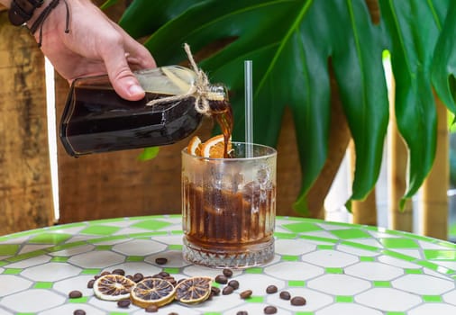 Hand pouring cold brew coffee in a bottle with transparent glass full of ice on green leaves background