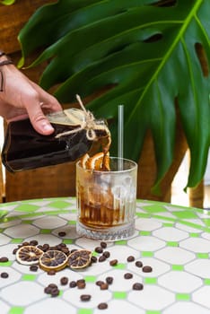 Hand pouring cold brew coffee in a bottle with transparent glass full of ice on green leaves background