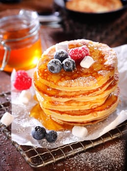 Stack of freshly baked pancakes drizzled with syrup or honey and topped with blueberries and raspberries with sprinkled sugar
