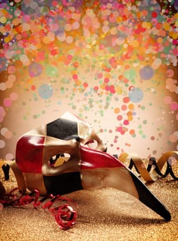 Close up Long Beak Carnival Venetian Masquerade Mask in Red Brown and Black Color with Paper Streamers on Glittery Table, Designed with Colorful Small Round Shapes on Brown Background.