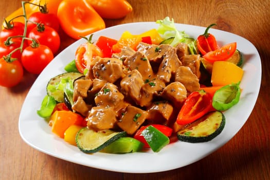 Close up Gourmet Saucy Cooked Meat or goulash with Fresh Veggies and Spices on a White Plate Served on Brown Wooden Table.