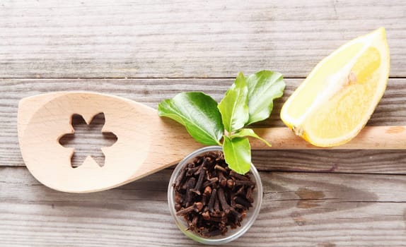 Overhead view of a decorative wooden spoon with a flower pattern cut out alongside a dish of dried aromatic cloves and a wedge of lemon on grey wooden rustic boards