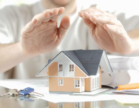 House Protection Concept - Bare Hands Over Miniature Model Home on Blueprint Placed on Top of Glass Table with Keys on the Side.