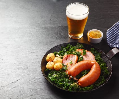Close up Appetizing German Recipe with Sausage, Pork Meat, and Potatoes on Green Vegetables, Served on a Frying Pan with Beer and Sauce on the Side. Dine in Black Wooden Table.