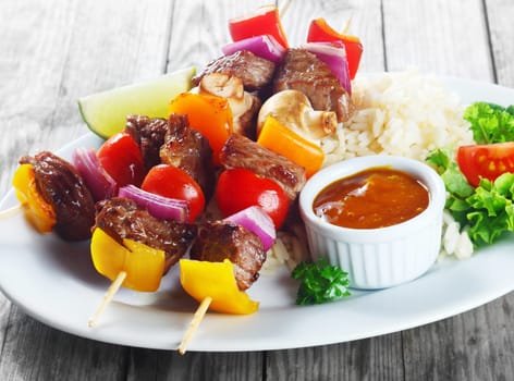 Gourmet Appetizing Kebab on Sticks with Sauce on White Plate with Rice and Veggies. Placed on Gray Wooden Table.