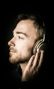 Attractive bearded young man musing as he listens to his music on stereo headphones looking off to the left in profile with a serene dreamy expression, close up of his face on a dark background