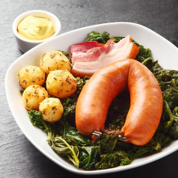 Close up Gourmet Cooked Sausage, Pork Meat and Potatoes on Green Leafy Vegetables, Served on White Plate with Sauce on the Side.