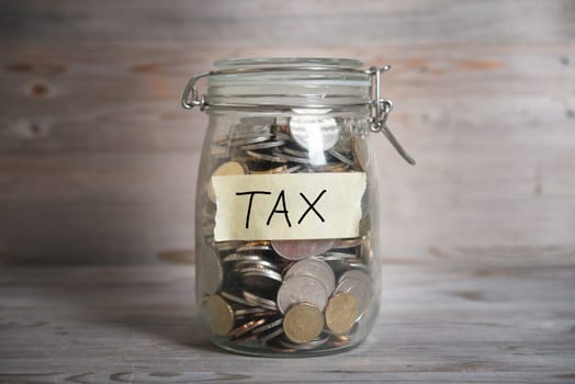 Coins in glass money jar with tax label, financial concept. Vintage wooden background with dramatic light.