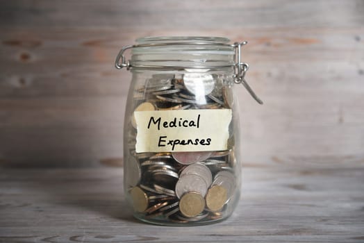 Coins in glass money jar with medical expenses label, financial concept. Vintage wooden background with dramatic light.
