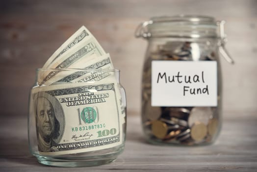 Dollars and coins in glass jar with mutual fund label, financial concept. Vintage tone wooden background with dramatic light.