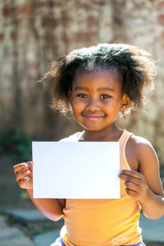 Portrait of little African girl showing blank white card outdoors.