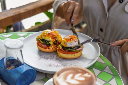 Hands with knife and fork cutting english muffin with poached eggs, bacon and Hollandaise sauce with vegetables