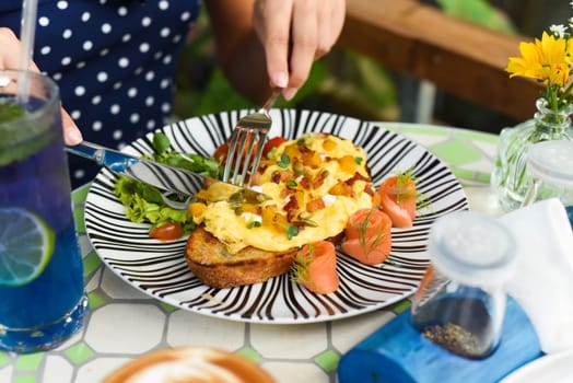 Woman holding knife and fork cutting toast with scrambled eggs pumpkin puree and cubes, smoked salmon and pumpkin seeds