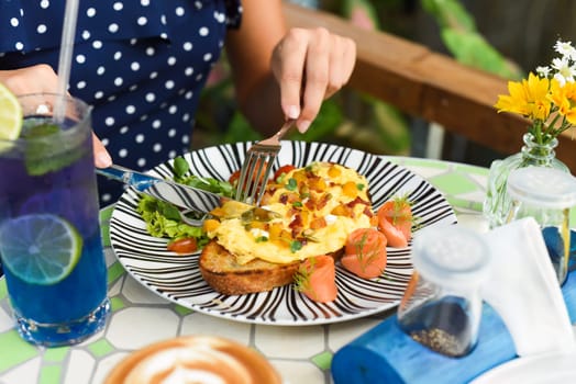 Woman holding knife and fork cutting toast with scrambled eggs pumpkin puree and cubes, smoked salmon and pumpkin seeds