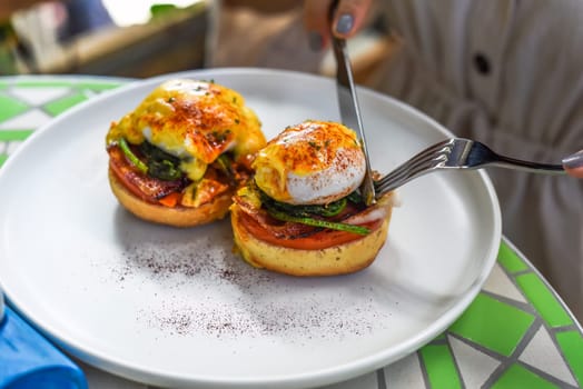 Hands with knife and fork cutting english muffin with poached eggs, bacon and Hollandaise sauce with vegetables