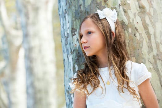 Portrait of young girl in white dress standing next to tree.