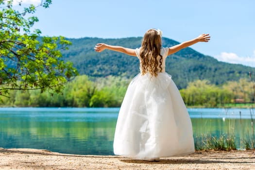 Young girl in white communion dress with open arms at lake.
