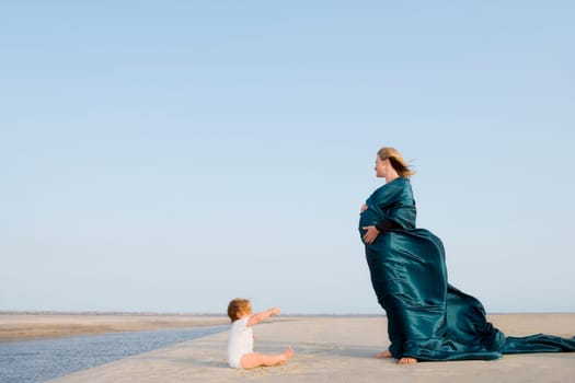 Side profile of a pregnant woman standing on the beach with her baby girl sitting in front of her