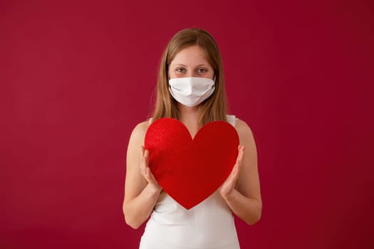 Smiling woman wearing face mask and holding red heart in hands. Concept of valentine during coronavirus pandemic. Cheerful blonde Caucasian girl from front view.