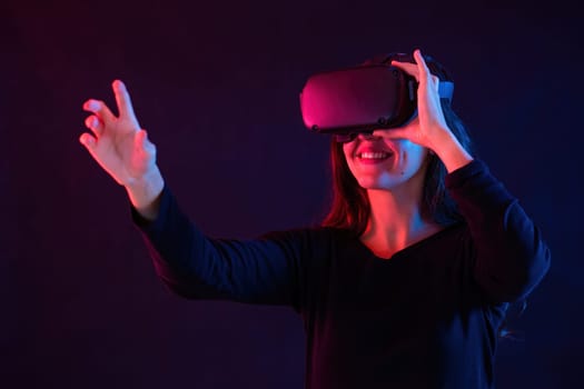 Smiling woman with headset interacting in virtual reality. Brunette using head equipment in dark space with red and blue light. Female touching fictitious thing.