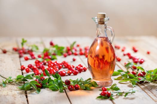 Bottle of rosehip oil on wooden palette with berries around. Clear oil fluid from sweet red fruit in jar with cork. Therapy with alternative medicine.