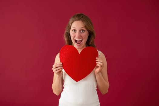 Excited Caucasian female with sparkling heart on valentines day. Happy woman holding love symbol on red background. Blonde girl full of happiness.