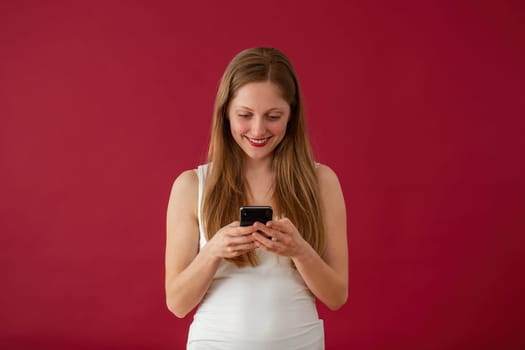 Smiling woman typing on mobilephone on red background. Blonde girl sending love on distance through the phone. Happy female reading message on smartphone.